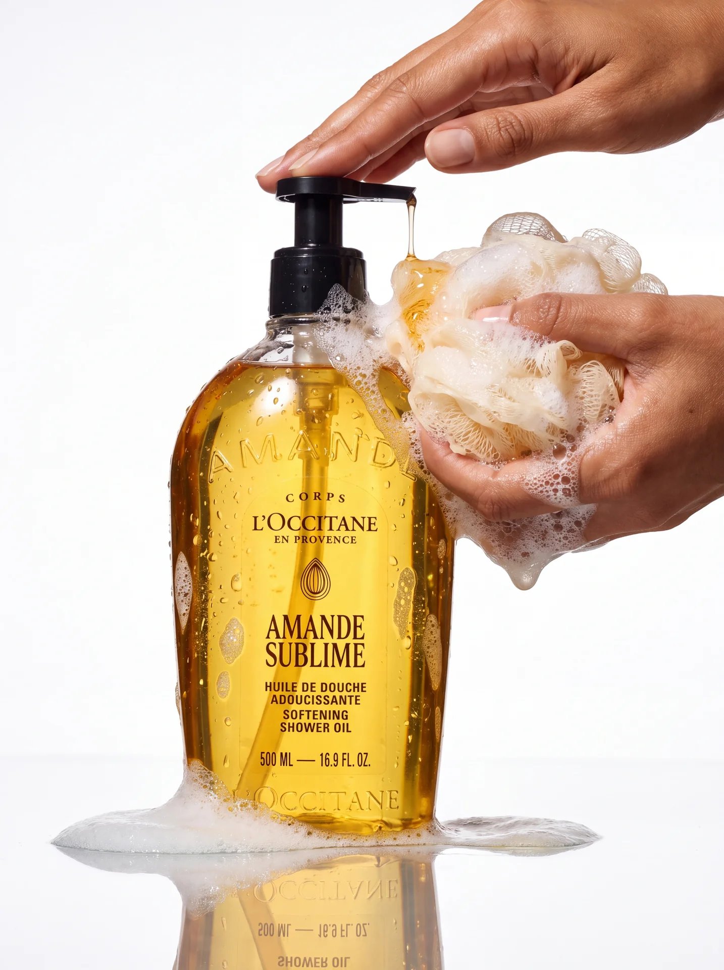 Hand lathering a sponge beside an Amande Sublime bottle on a mirrored surface