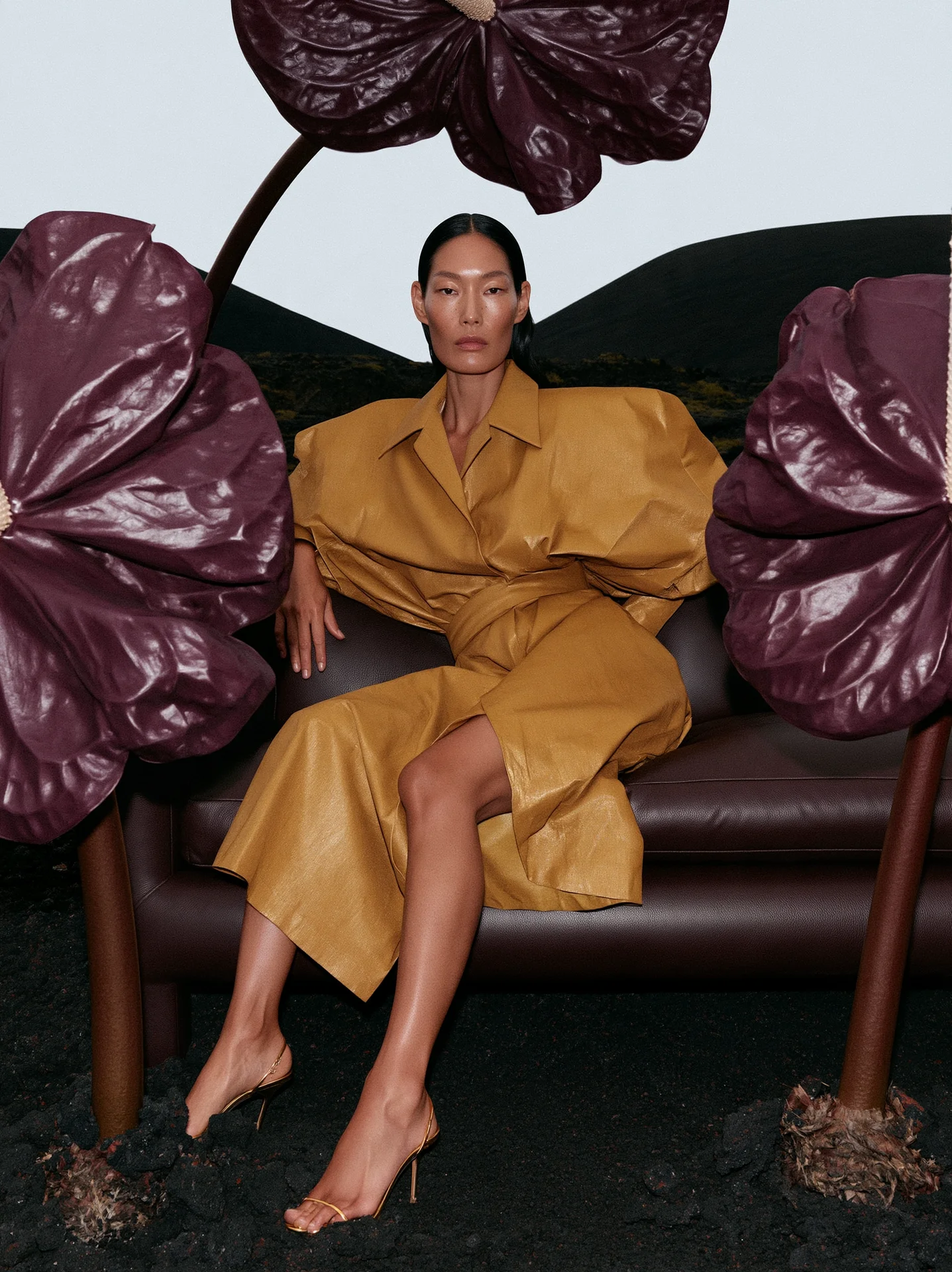 Mustard-yellow leather coat seated on a brown leather sofa beside giant dark-purple leaves