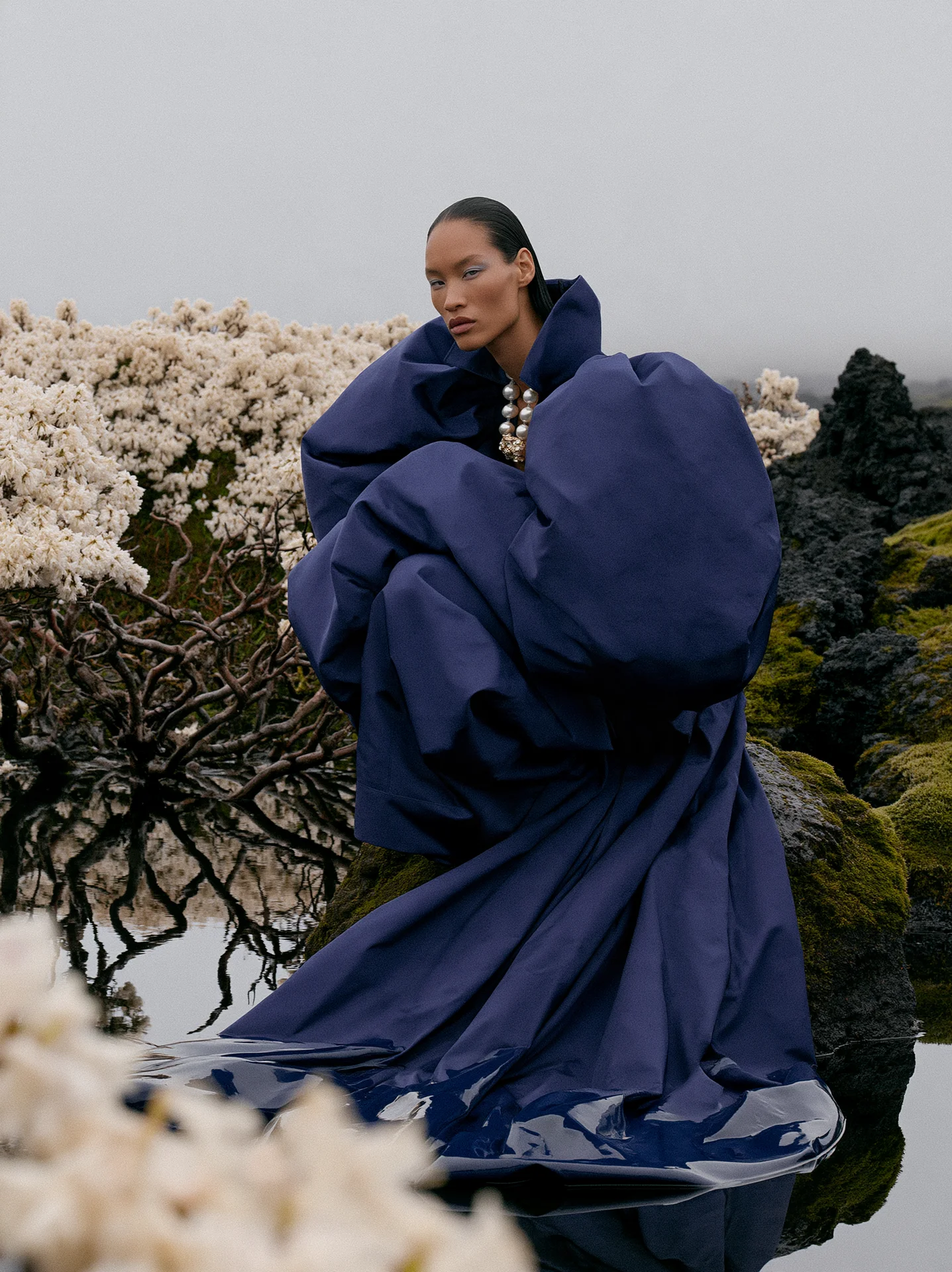 Navy-blue sculptural rose gown among white blossom branches with mountains behind