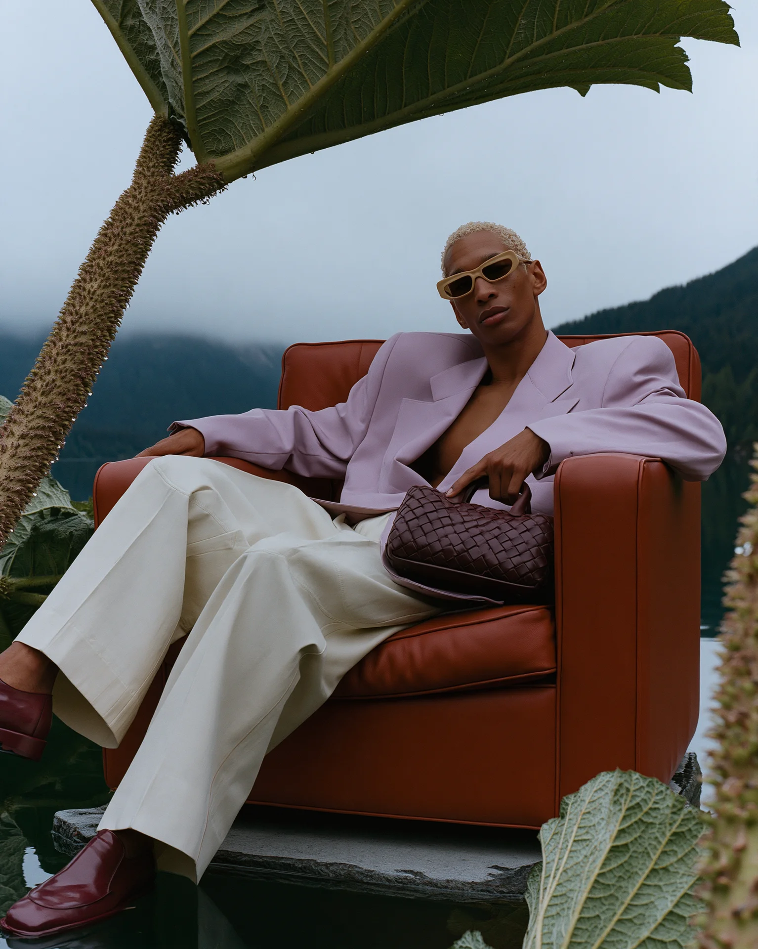 Man in a lavender suit seated in an orange armchair beneath giant tropical leaves