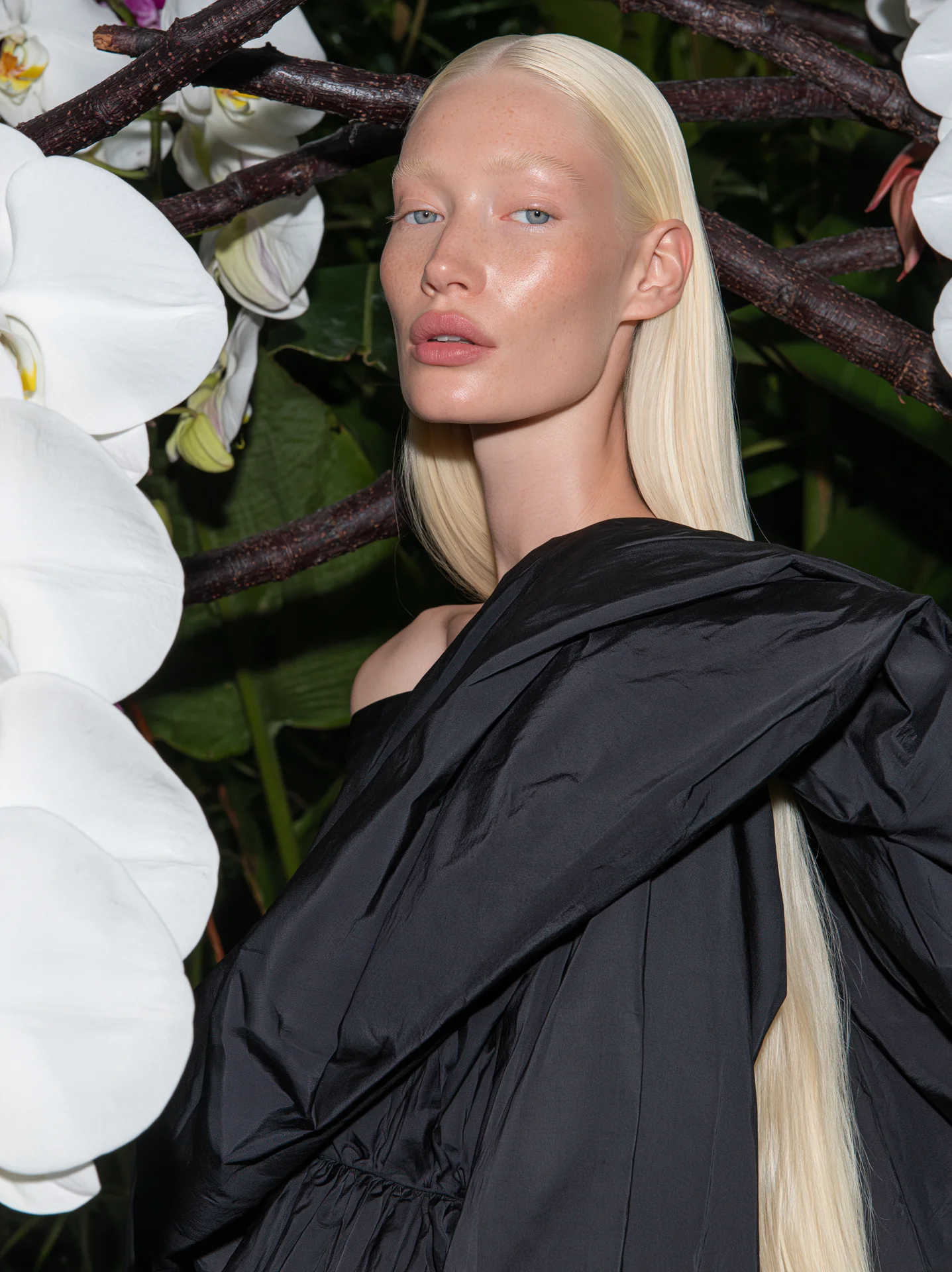 Platinum-blonde model in a black draped garment beneath a dark tree with white orchids