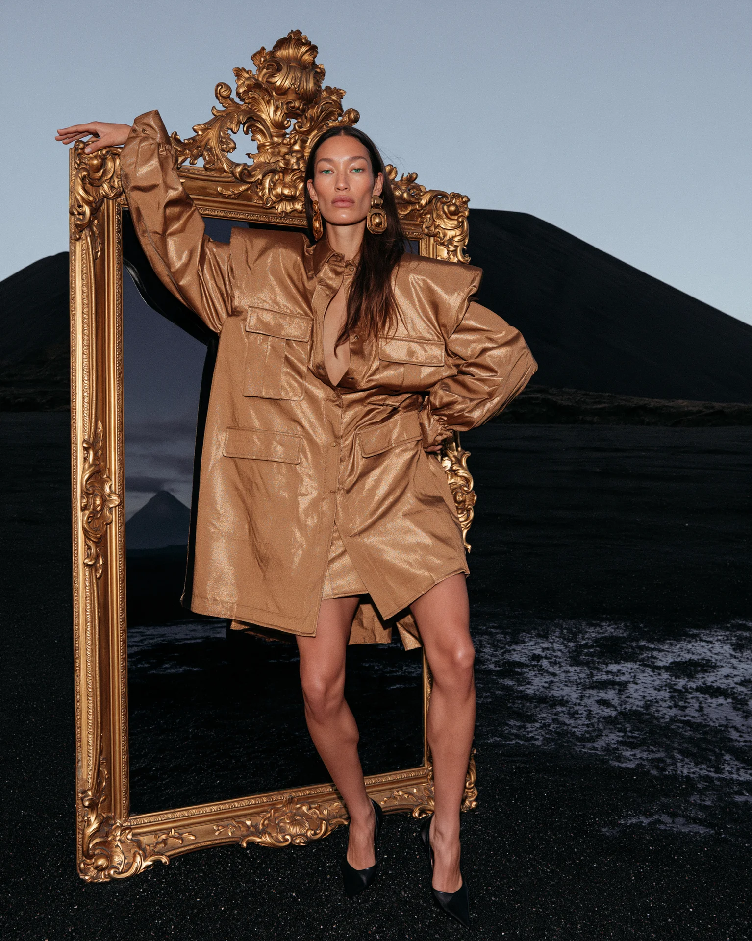 Copper-brown dress, figure standing inside a giant ornate gold picture frame on a black-sand shore